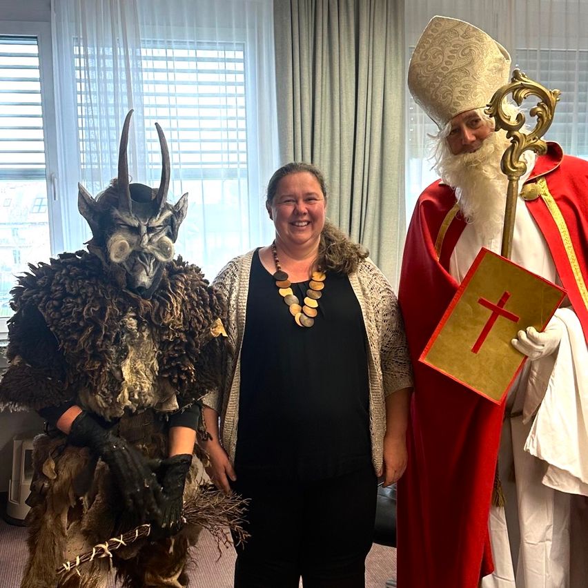 Eine Frau in einem schwarzen Kleid lächelt für ein Foto mit einem Mann in einem hörnertragenden Teufelskostüm und einem Mann in einem roten Bischofskostüm.