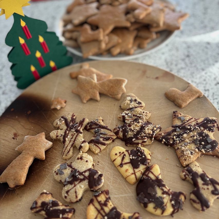 Ein Holzschneidebrett zeigt verschiedene mit Schokolade überzogene und dekorierte Kekse, mit einem Hintergrund von weiteren Keksen auf einem Teller und einer ausgeschnittenen Weihnachtsbaumdekoration.