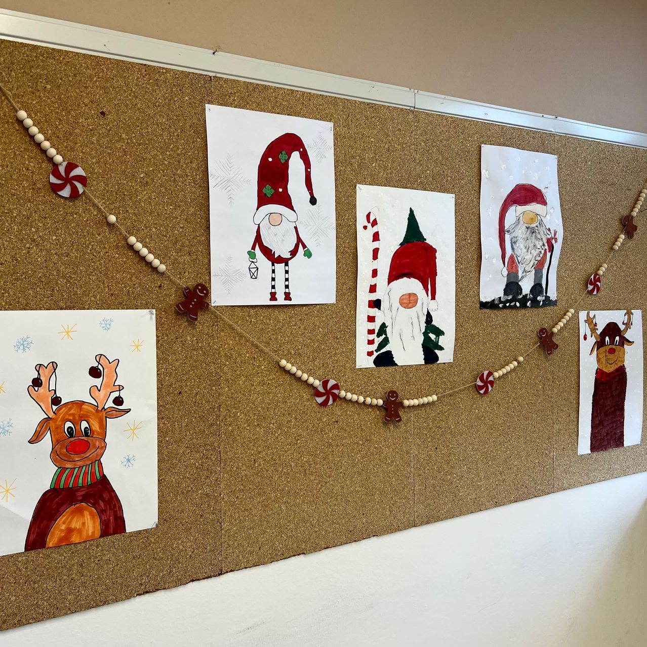 Eine Korktafel zeigt verschiedene weihnachtliche Zeichnungen, darunter Weihnachtsmann, Lebkuchenmann und Rentier, zusammen mit einer dekorativen Girlande.