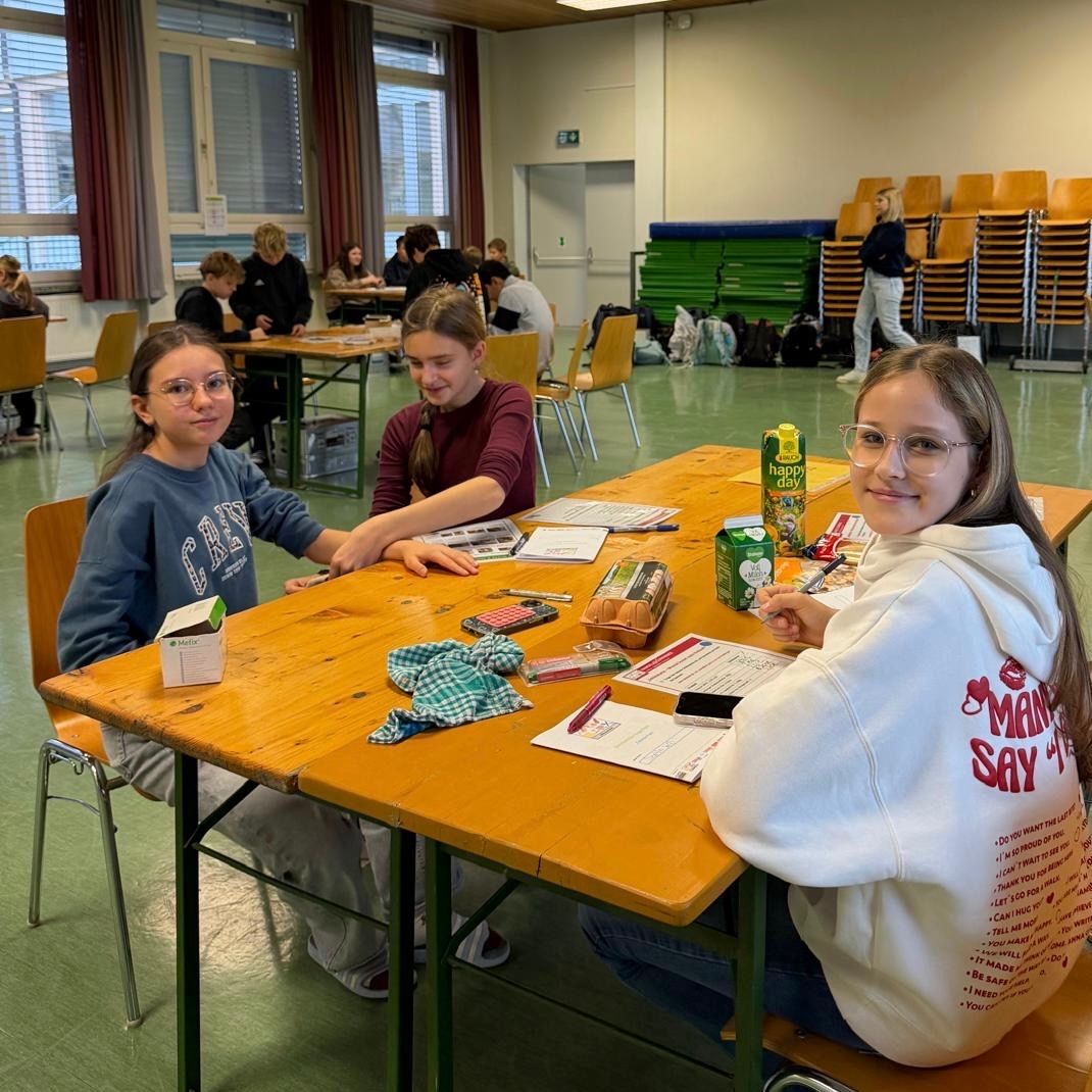 Drei Mädchen mit Brille sitzen lächelnd und interagieren an einem Tisch. Sie haben eine Saftbox, ein Sandwich und verschiedene Gegenstände auf dem Tisch. Hinter ihnen sitzen weitere Schüler an Tischen, und eine Person geht im Hintergrund vorbei.