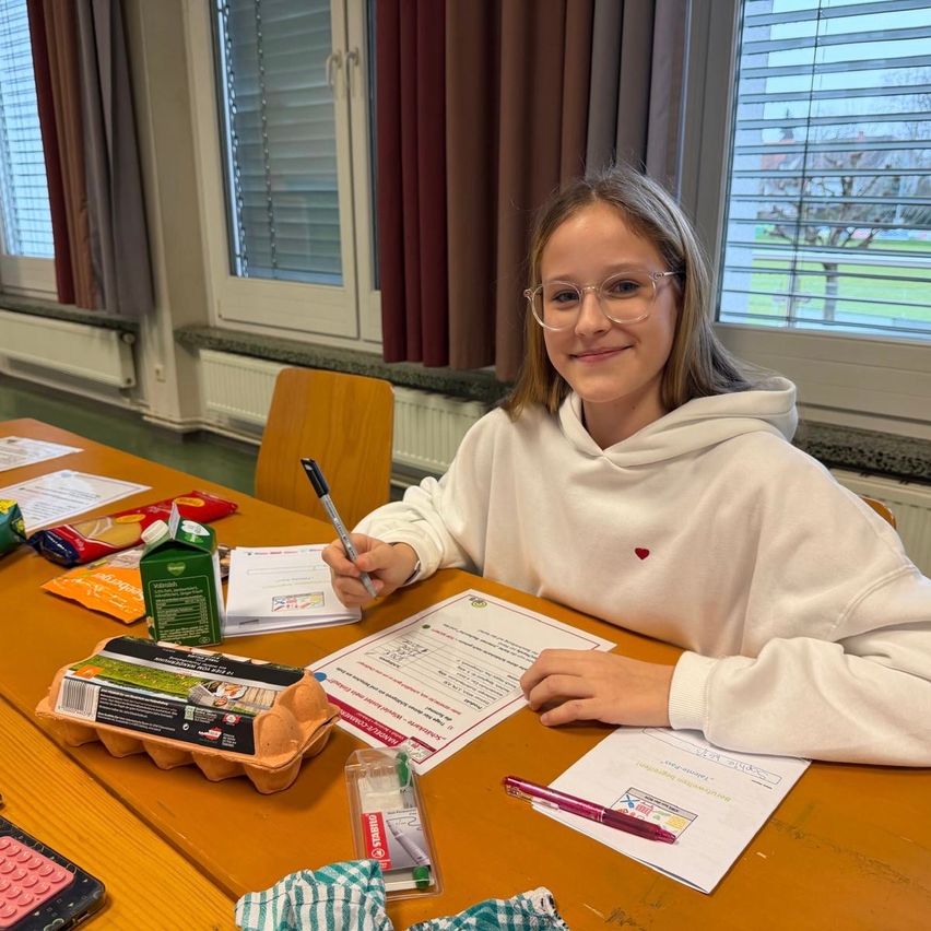 Ein junges Mädchen in einem weißen Hoodie sitzt mit einem Lächeln am Tisch und schreibt. Auf dem Tisch liegen Papiere, ein Taschenrechner und andere Gegenstände. Hinter ihr befinden sich Fenster mit braunen Vorhängen.