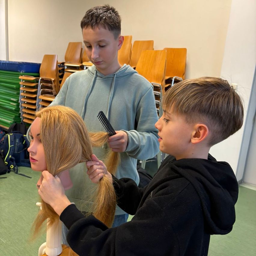 Ein junger Junge übt das Friseurhandwerk an einer Puppe, während ein anderer Junge zuschaut, mit Reihen von Stühlen im Hintergrund.