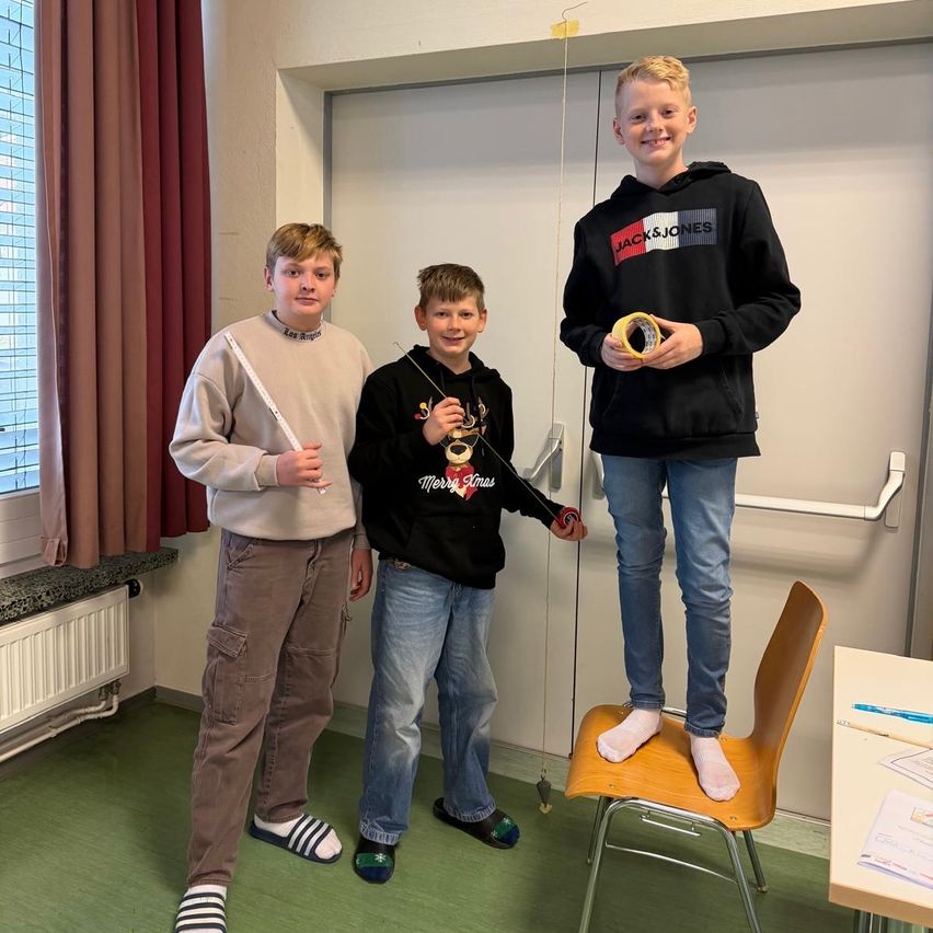 Drei Jungen in einem Raum. Einer steht auf einem Stuhl, ein anderer hält eine Angelrute und der dritte ein Klebeband.