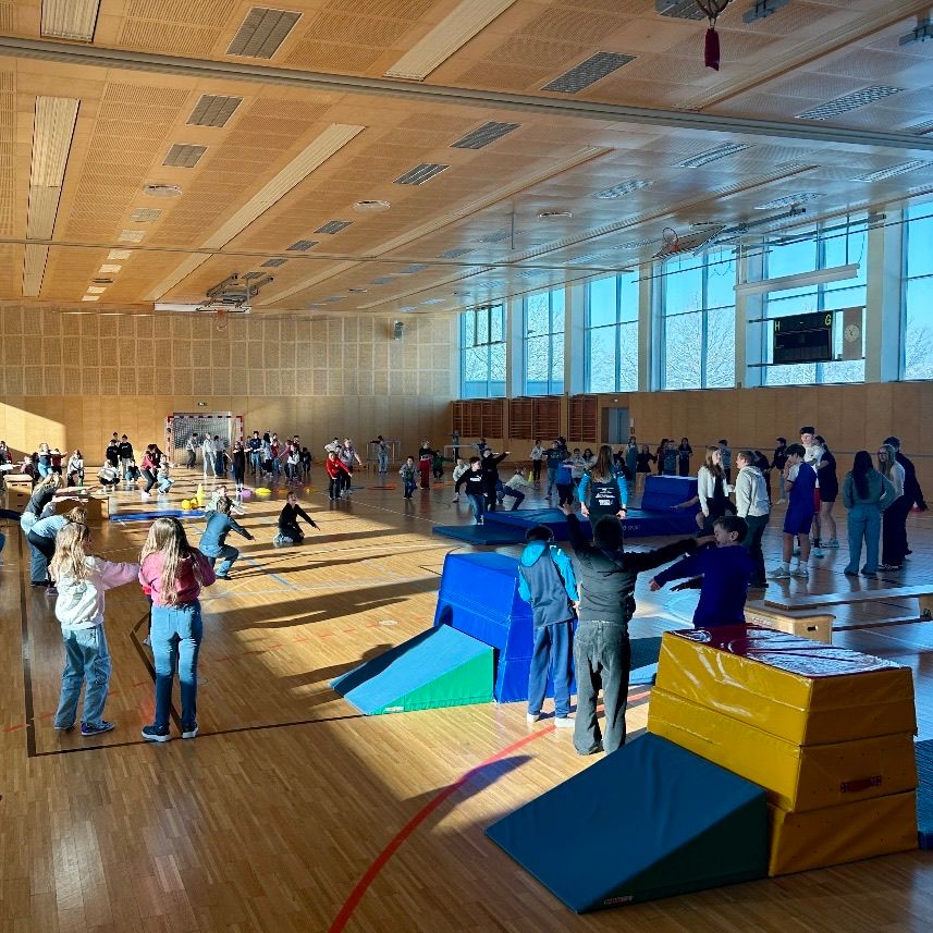 Ein Sportsaal voller spielender Kinder, einige auf verschiedenen farbigen Matten und Hindernissen, andere stehend und zuschauend. Die Wände sind aus Holz, mit großen Fenstern, die Sonnenlicht hereinlassen.