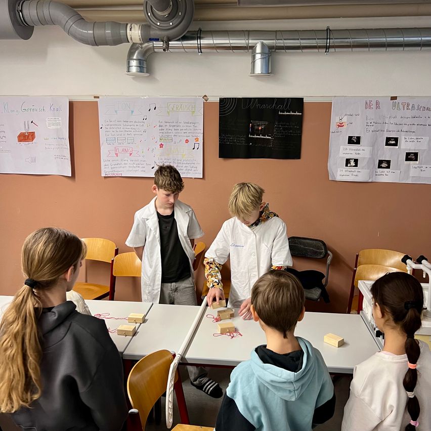 Eine Gruppe von Schülern in Laborkitteln versammelt sich um einen Tisch mit Objekten. Sie scheinen an einem wissenschaftlichen Experiment beteiligt zu sein. Der Raum hat Poster und eine Wand mit angehängten Objekten.