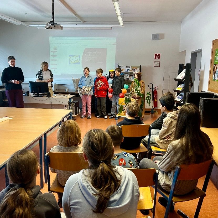 In einem Klassenzimmer stehen mehrere Schüler vor einer Projektionsleinwand, während andere auf Stühlen sitzen. Zwei Erwachsene stehen in der Nähe. Das Klassenzimmer ist gut beleuchtet, und es sind Tische, Bücher und ein Feuerlöscher sichtbar.