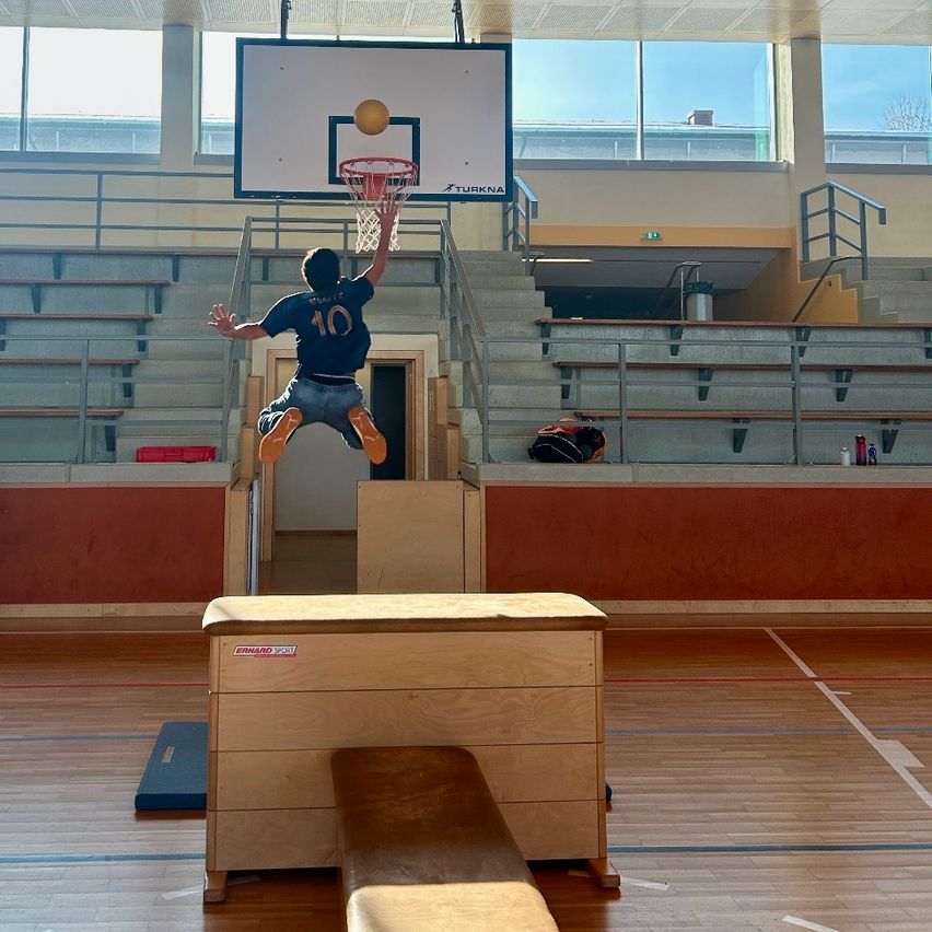 Ein junger Mann springt, um einen Basketballkorb in einer Indoor-Sporthalle zu dunken. Die Tribünen sind leer, und eine Tasche liegt auf dem Boden.