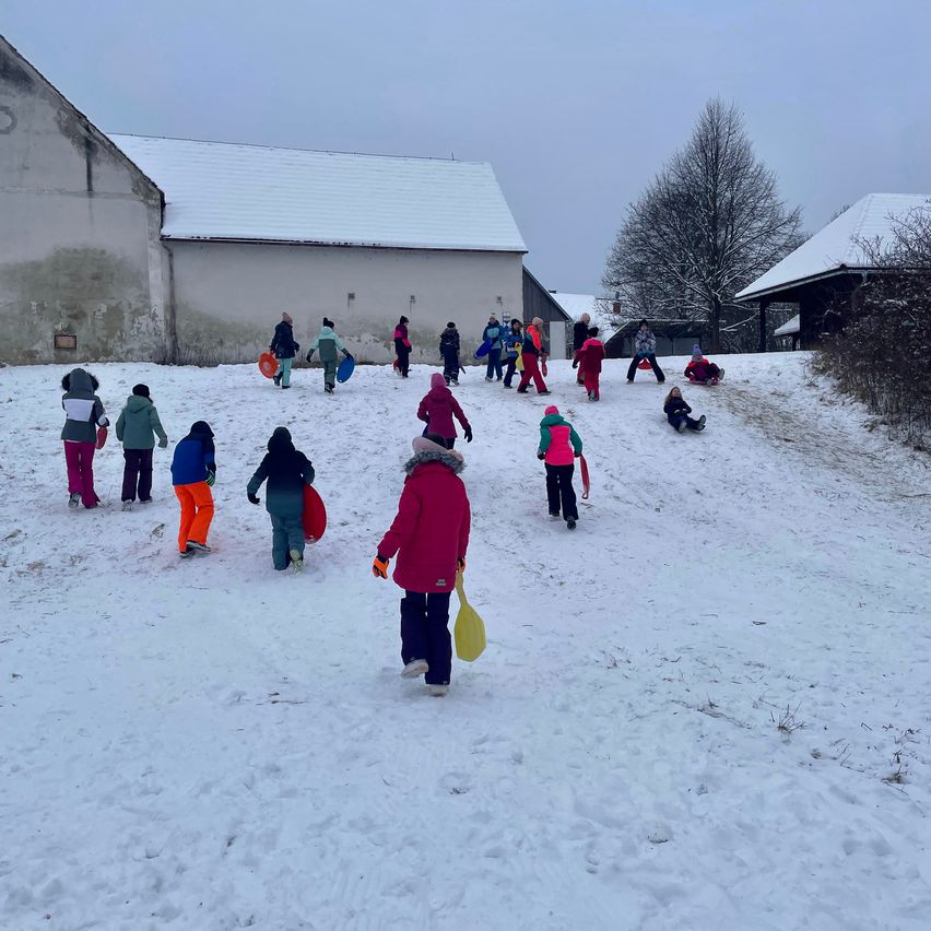 Kinder in bunter Winterkleidung spielen im Schnee, einige mit Schlitten und andere gehen einen schneebedeckten Hügel hinauf. Im Hintergrund befinden sich Gebäude mit schneebedeckten Dächern und ein kahler Baum.