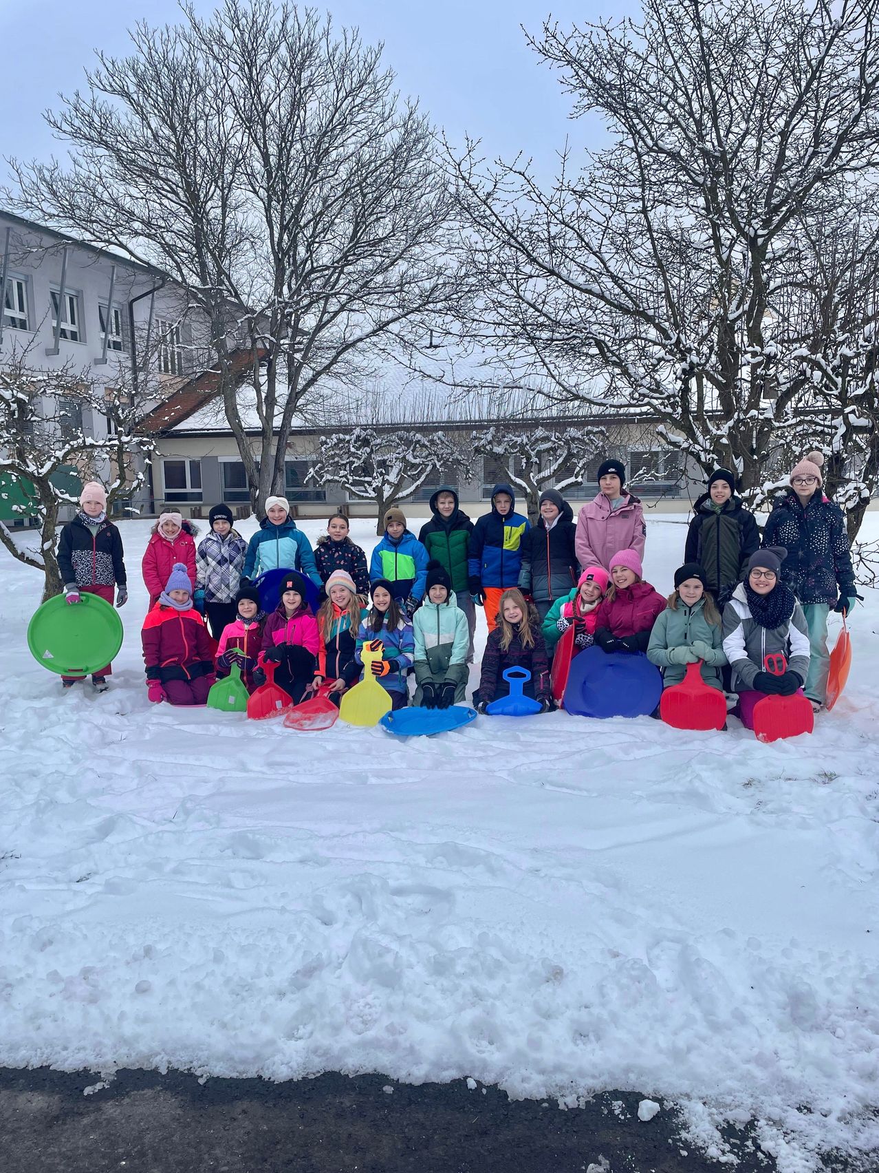 Eine Gruppe von Kindern und Erwachsenen posiert für ein Foto im Schnee mit bunten Schneeschaufeln vor einem Gebäude mit Bäumen.