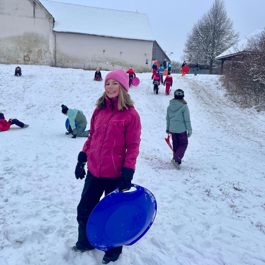 Ein Mädchen in einer rosa Jacke steht auf einem verschneiten Hügel mit einem blauen Schlitten, während andere Kinder hinter ihr im Schnee spielen.