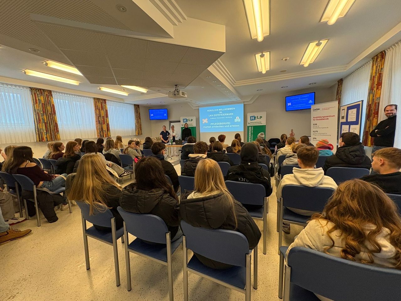 A classroom with a projector screen showing a presentation, several people seated, and two individuals standing in front of the screen. The room has blue chairs, a white ceiling with lights, and two monitors mounted on the wall.