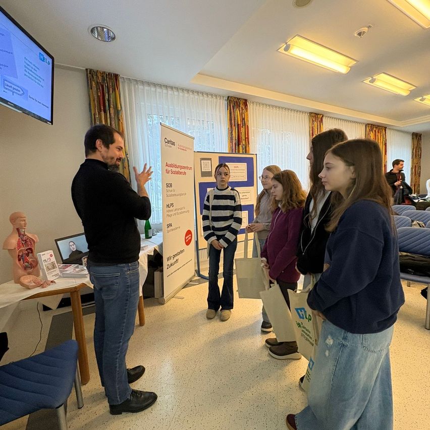 A man stands next to a display, presenting to a group of students in a classroom. A mannequin and books are on a table behind him. The students listen and some hold bags.