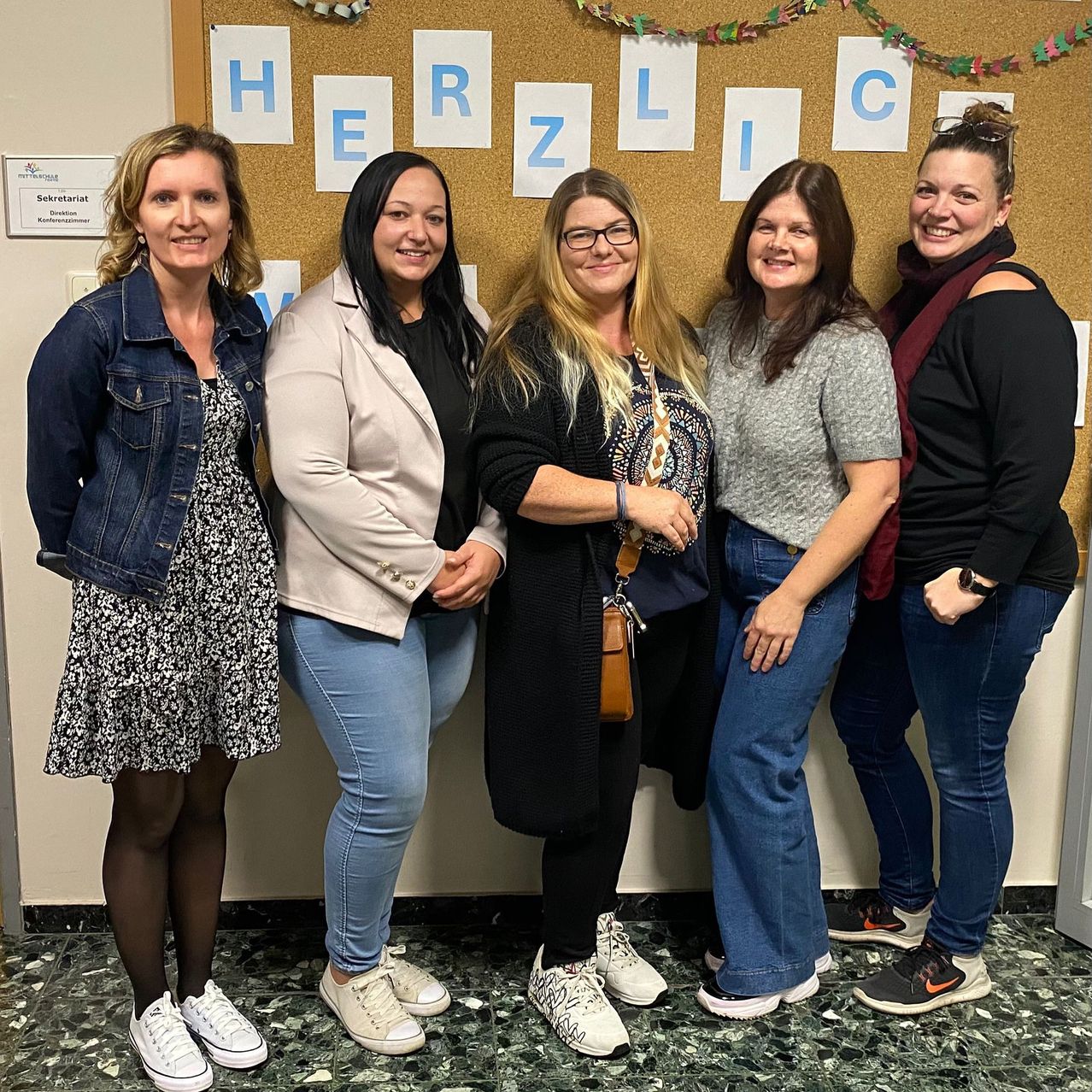Fünf Frauen stehen lächelnd zusammen vor einem Aushang mit den Buchstaben H, E, R, Z, L, I und C.