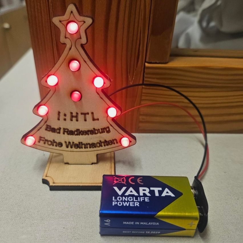 Ein holzern Weihnachtsbaum mit roten LED-Lichtern ist an eine Batterie angeschlossen. Der Baum hat die Worte 'Bad Rackersburg Frohe Weihnachten' darauf geschrieben. Eine Batterie mit der Aufschrift 'VARTA LONGLIFE POWER' steht daneben.