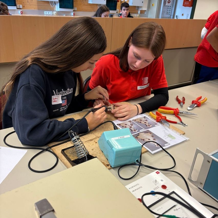 Zwei Mädchen arbeiten in einer Werkstatt an einem Lötprojekt. Eine trägt ein Manhattan-Sweatshirt, die andere ein rotes Hemd. Werkzeuge und Papiere umgeben sie.