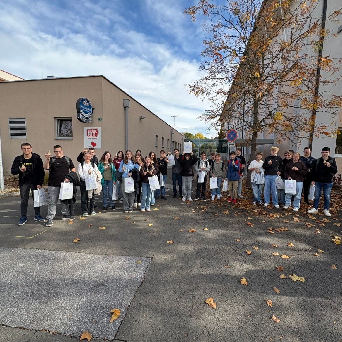 Eine Gruppe von Menschen, möglicherweise Schüler, steht an einem sonnigen Tag in einer Reihe vor einem Gebäude. Sie halten alle weiße Taschen und scheinen für ein Foto zu posieren.