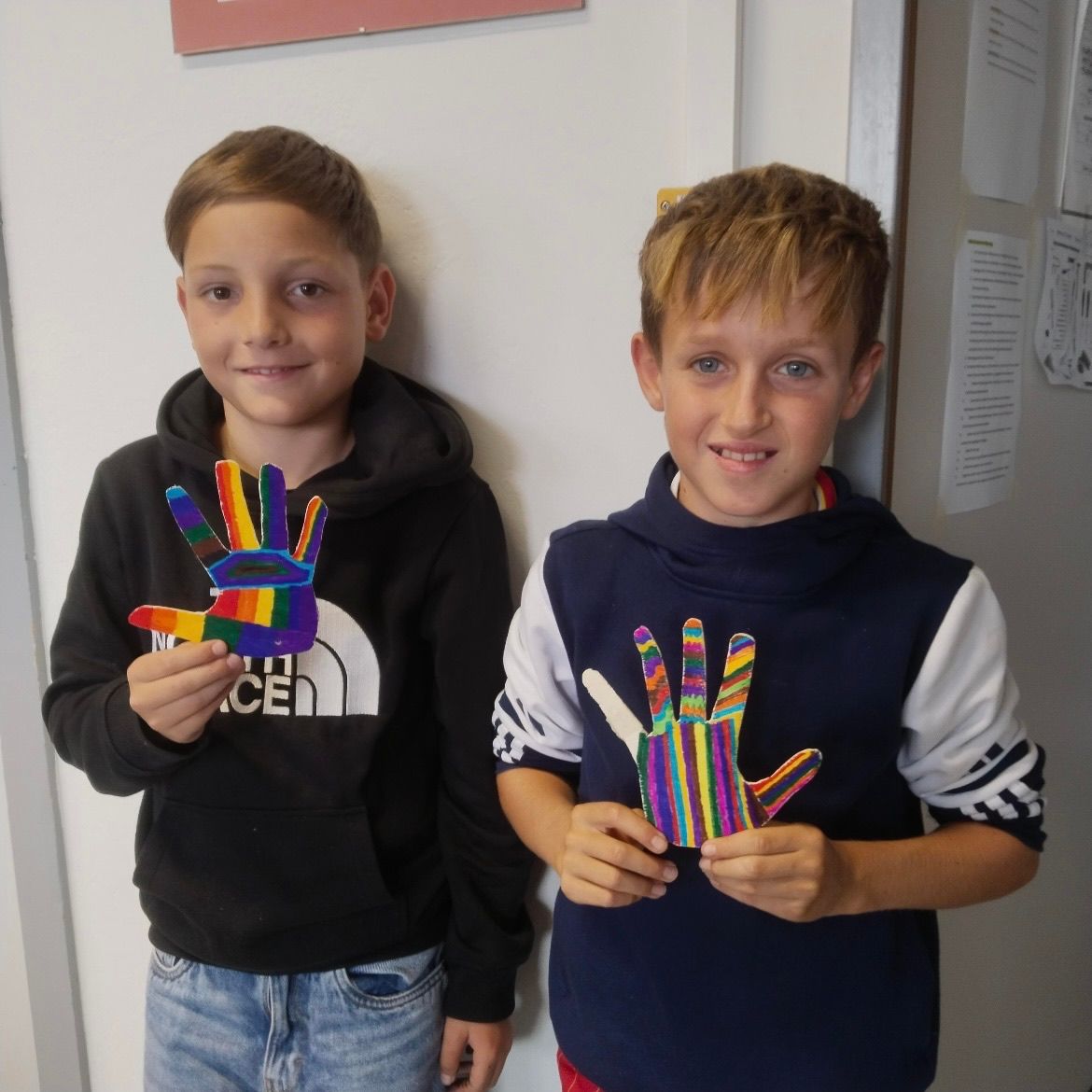 Zwei junge Jungen stehen nebeneinander und halten jeweils eine farbenfrohe Papierhand. Ein Junge trägt eine schwarze Kapuzenjacke, der andere ein blaues Weste.