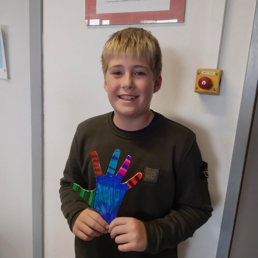 Ein Junge mit einer bemalten Hand hält sie hoch und lächelt, während er vor einer weißen Wand steht.