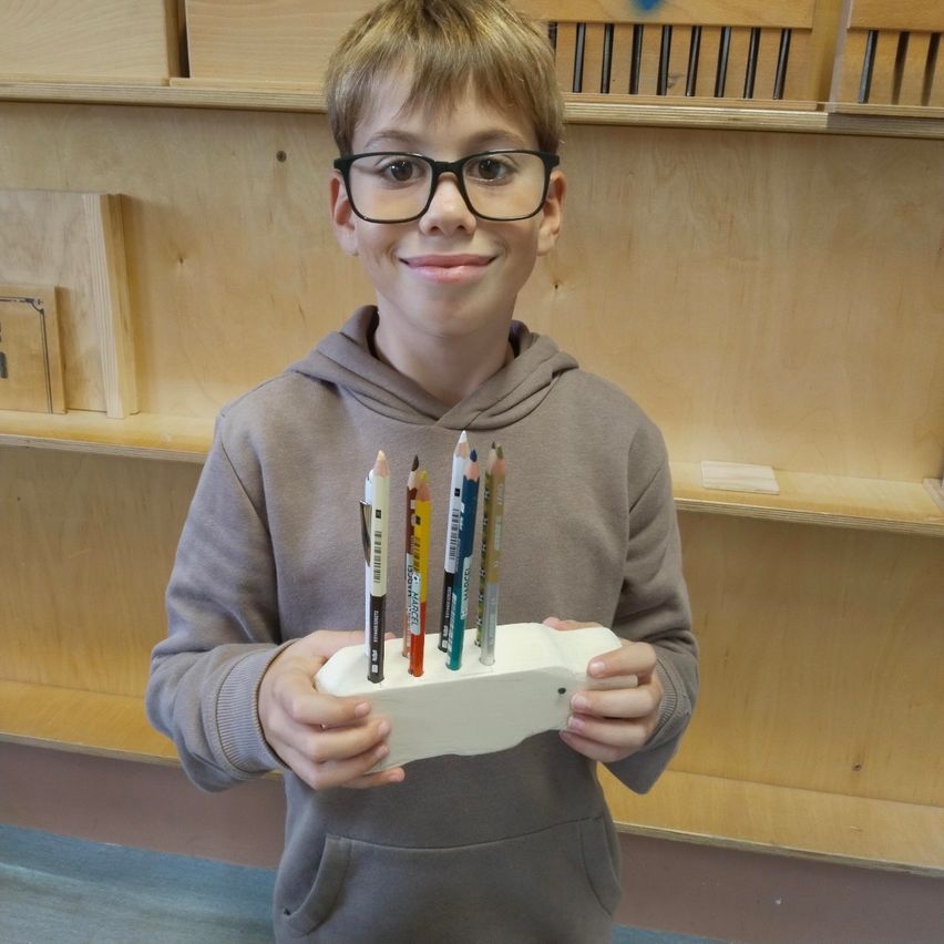 Ein junger Junge mit Brille und einer grauen Kapuzenjacke hält einen weißen Stifthalter mit darin angeordneten Bleistiften und steht vor einem Holzregal.