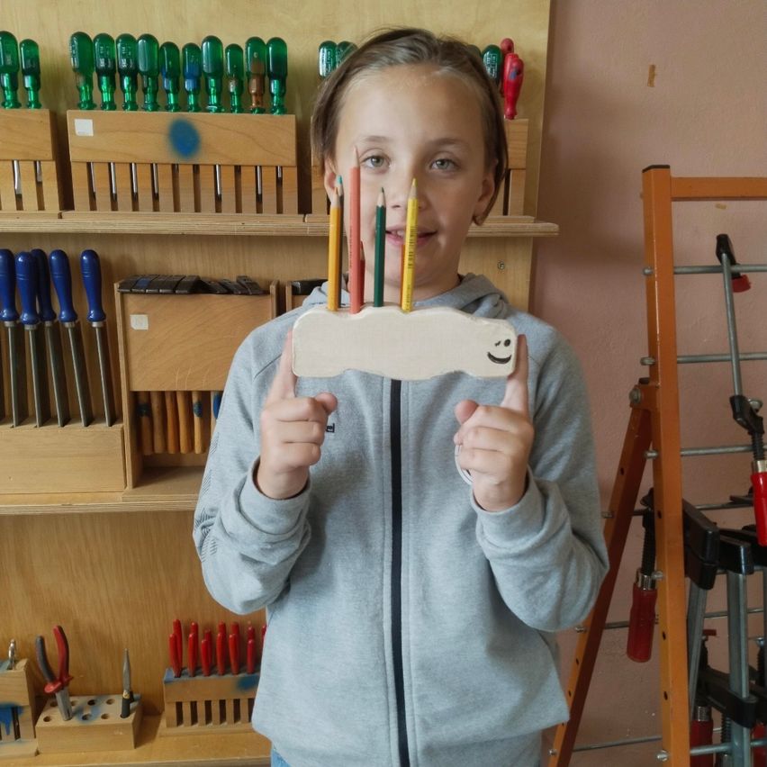 Ein junges Mädchen steht in einer Werkstatt und hält einen holzernen Stifthalter mit bunten Bleistiften. Hinter ihr steht ein Holzregal mit verschiedenen Werkzeugen.