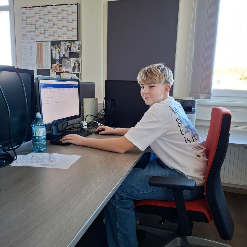 Ein Junge sitzt in einem Bürostuhl vor einem Schreibtisch mit einem Computer und einer Wasserflasche. Er lächelt und scheint am Computer zu arbeiten.