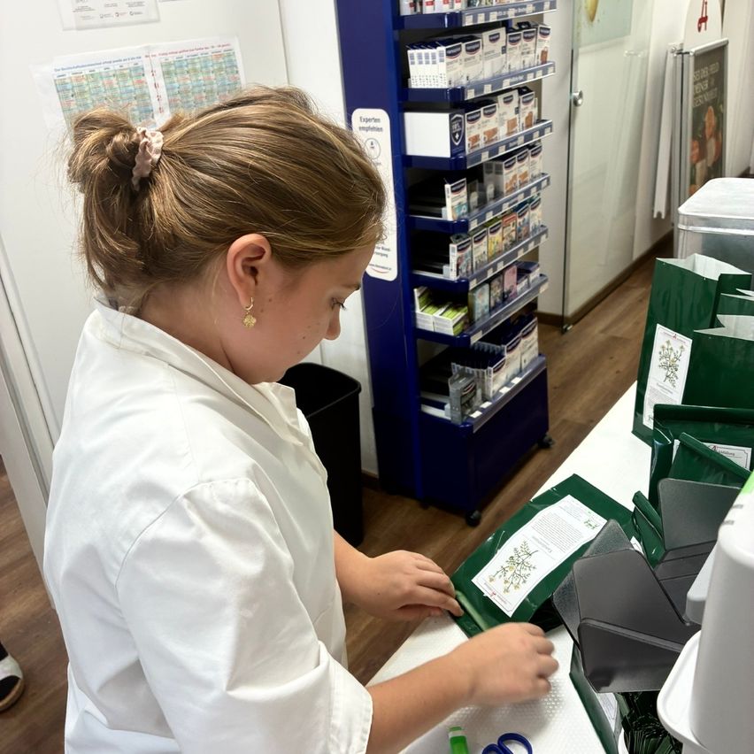 Eine Frau in einem weißen Laborkittel verpackt grüne Produkte auf einem Tresen. Hinter ihr befinden sich Regale mit verschiedenen Produkten.
