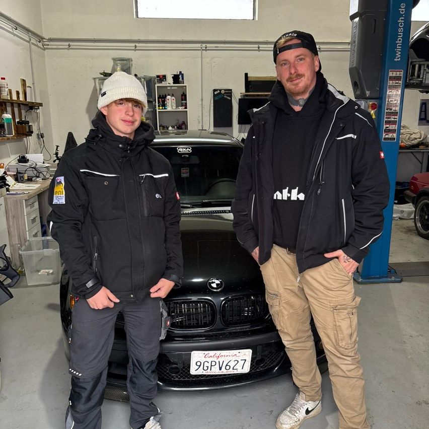 Zwei Männer in Winterjacken stehen in einer Garage und posieren vor einem schwarzen BMW mit einem kalifornischen Kennzeichen. Der Mann auf der linken Seite trägt eine Mütze und Turnschuhe. Hinter ihnen sind Regale und ein Autolift zu sehen.