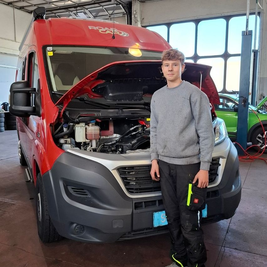 Ein junger Mann steht vor einem roten Van mit offenem Motorhaube in einer Garage. Er trägt ein graues Sweatshirt und schwarze Hosen.