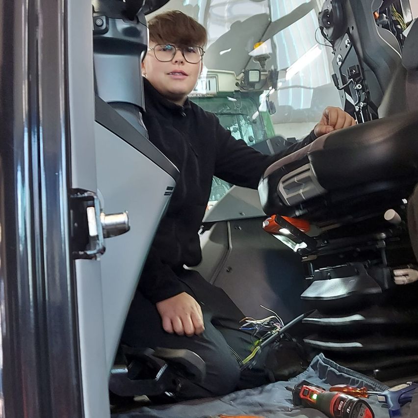 Eine junge Person sitzt in einem Fahrzeug, wahrscheinlich einem Traktor, und trägt eine Brille. Sie hat Werkzeuge auf dem Boden und lehnt sich gegen den Sitz.
