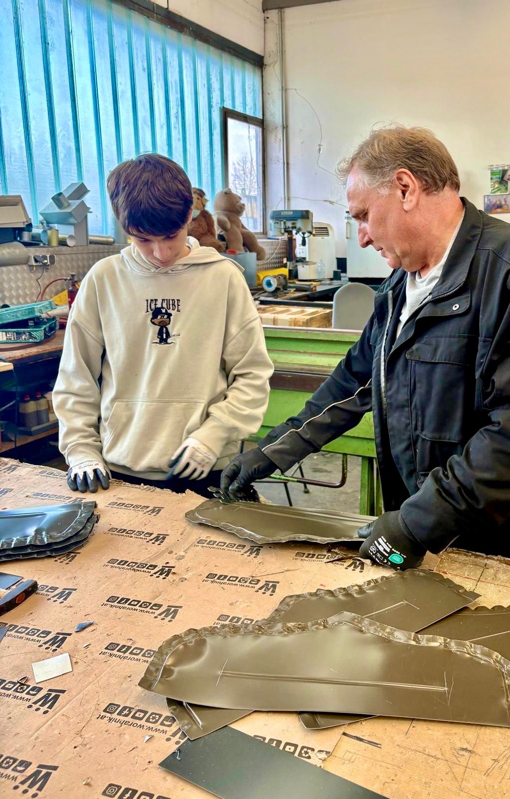 Ein älterer Mann und ein Junge arbeiten in einer Werkstatt an einem Metallprojekt. Der Junge trägt eine weiße Kapuzenjacke mit einem Grafikaufdruck. Der Mann trägt eine schwarze Jacke. Sie tragen beide Handschuhe. Vor ihnen steht ein Tisch mit Metallblechen darauf. Im Hintergrund befinden sich Werkzeuge und Maschinen.