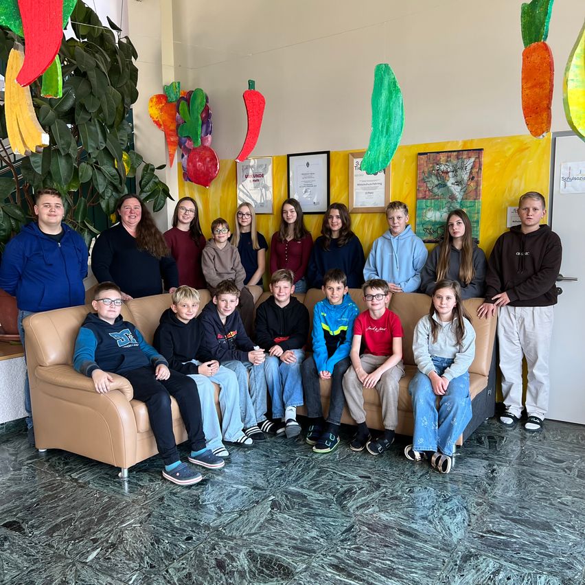 Eine Gruppe von Schülern und Lehrern posiert für ein Foto in einem Raum mit einer gelben Wand, die mit verschiedenen Kunstwerken und hängenden Gemüsestücken verziert ist.