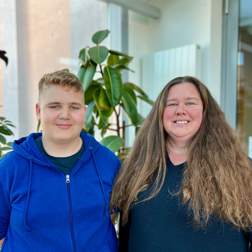 Zwei Personen stehen nah beieinander drinnen und lächeln für ein Foto. Ein Mann in einer blauen Kapuzenjacke und eine Frau in einem blauen Shirt stehen vor einer Glaswand.