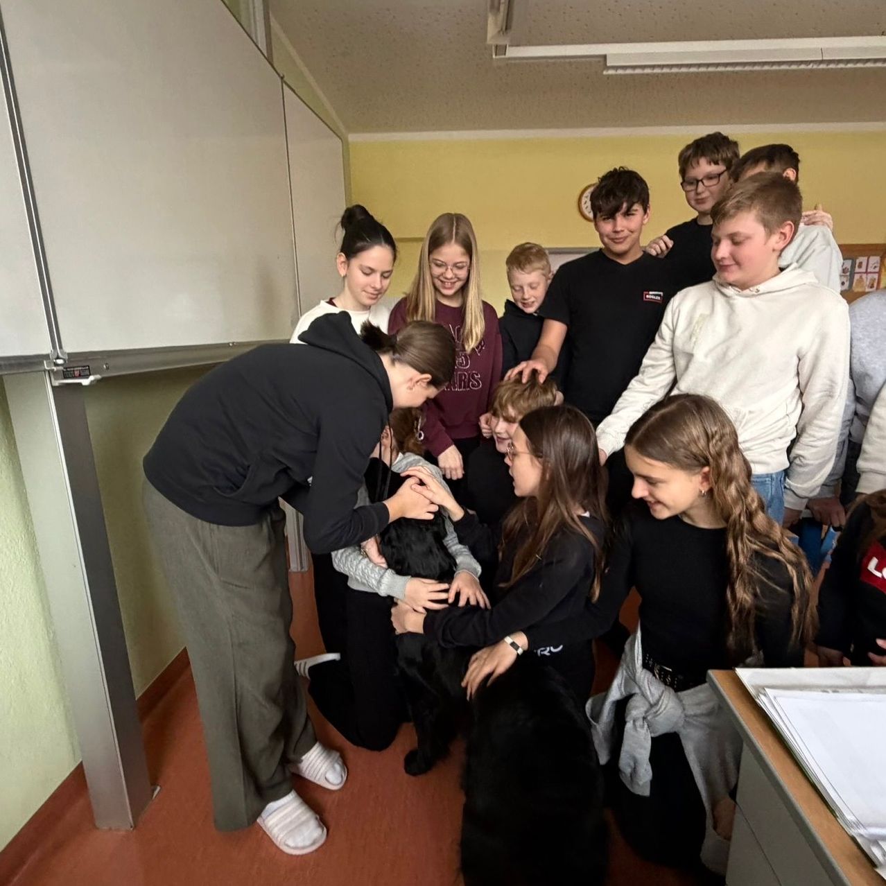 A group of young people are gathered in a room with a dog, some kneeling and some standing, all smiling and enjoying the dog's presence.