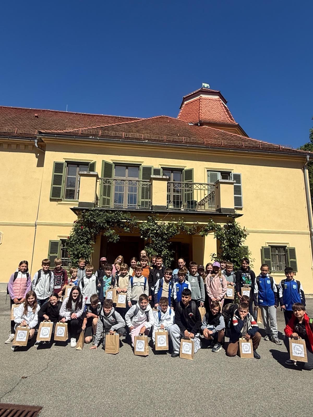 Eine Gruppe von Kindern und Jugendlichen in unterschiedlicher Kleidung posiert für ein Foto vor einem gelben Gebäude mit rotem Dach und Balkonen mit grünen Fensterläden.