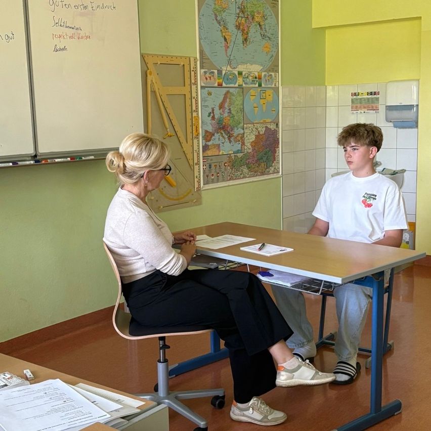 A teacher and a student sit at a desk in a classroom. A whiteboard and a world map are in the background.