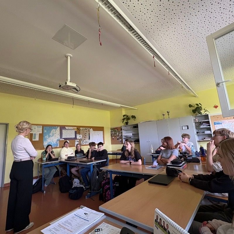 Classroom with yellow walls, a teacher speaking, and students seated in front of tables, with various items like backpacks and books on the desks.