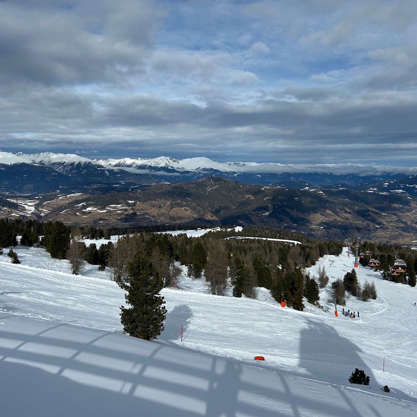 Aerial view of a snowy mountain landscape with pine trees and ski slopes. In the distance, a snow-covered mountain range is visible under a partly cloudy sky.
