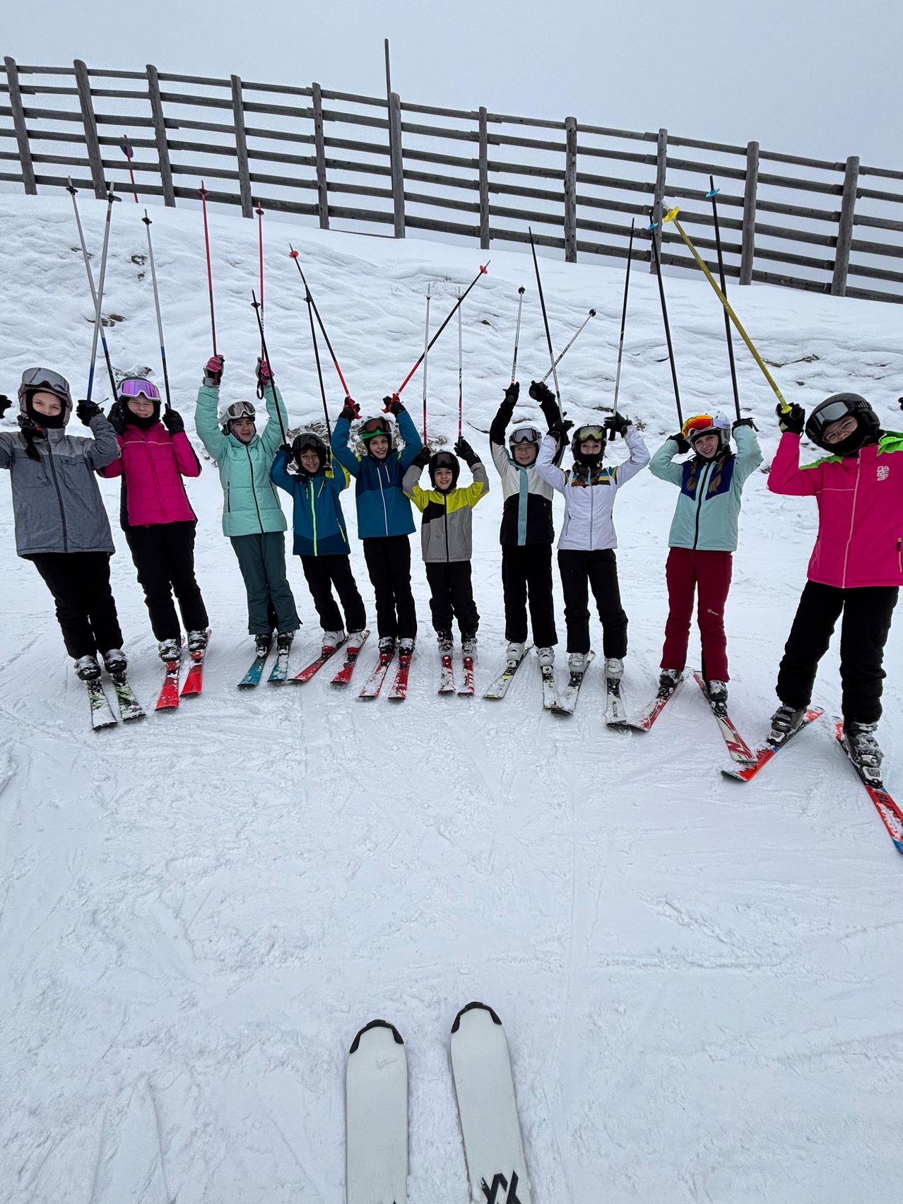 A group of skiers is posing for a photo on a snowy mountain. They are all dressed in ski gear and holding their ski poles. The sky is overcast.