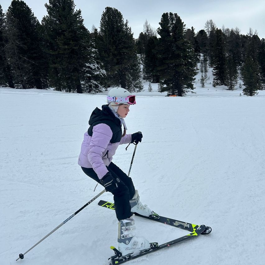 A young girl in winter gear skiing on a snowy slope, surrounded by evergreen trees.