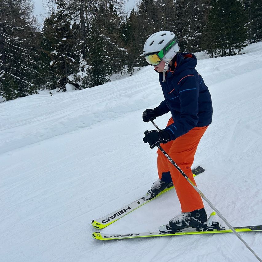 A skier wearing an Atomic helmet and goggles is skiing down a snowy slope. They are dressed in a blue jacket, orange pants, and black gloves. The skis are yellow and black.