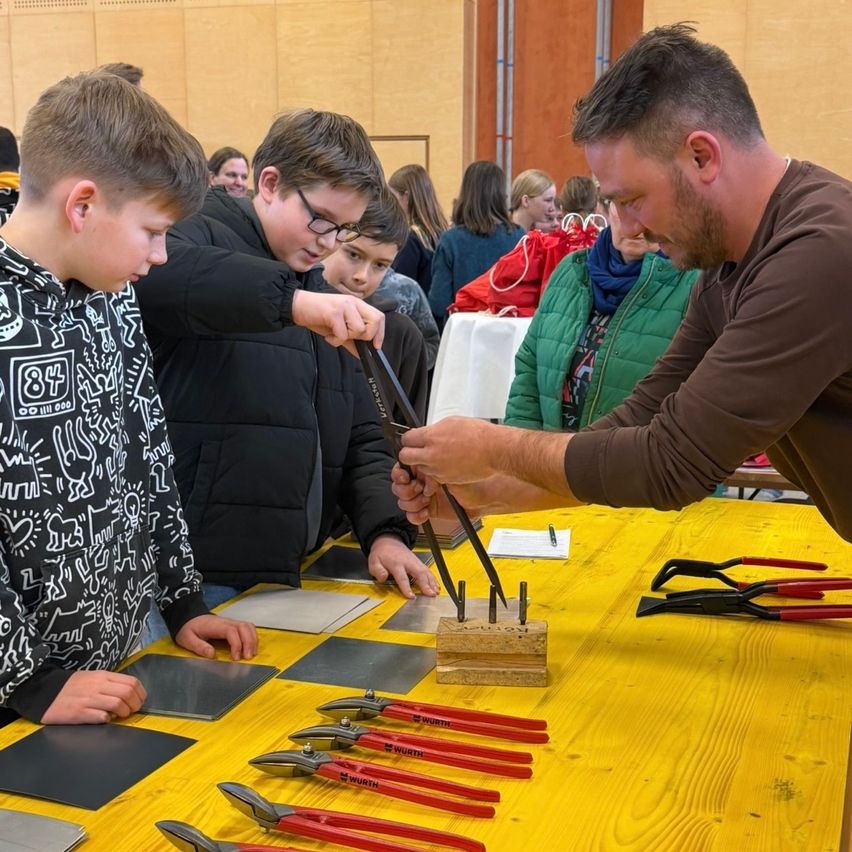 A man demonstrates metalworking to two boys while several others watch, inside a room with wooden walls.