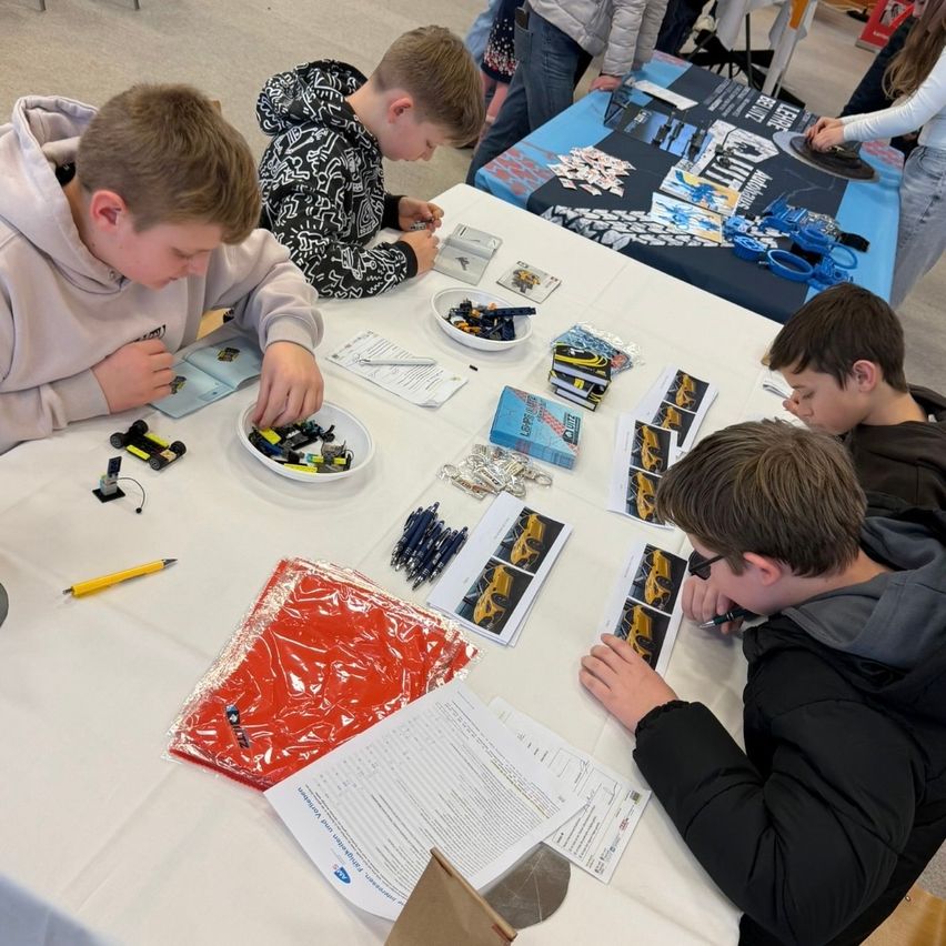 Several boys are gathered around a table with a variety of toys and tools. One boy looks at a sheet of paper while another holds a pencil. The table is covered with a white tablecloth.