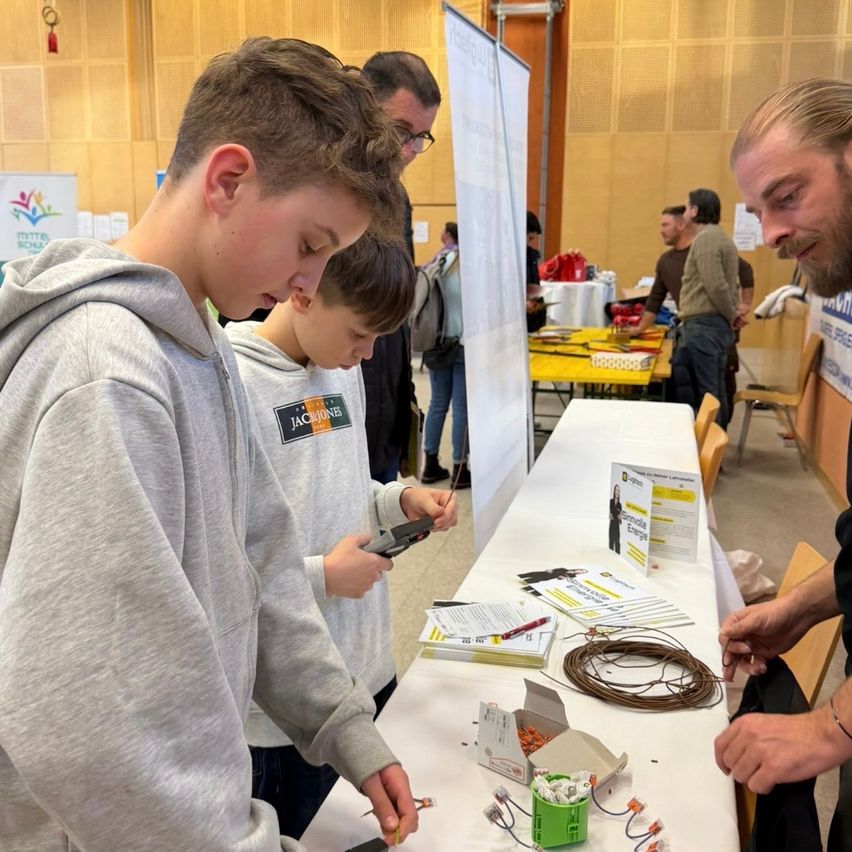 Two boys are working on an electrical project at a table with a man, possibly a teacher, guiding them. The table has various materials. Behind them, a person stands by a banner.