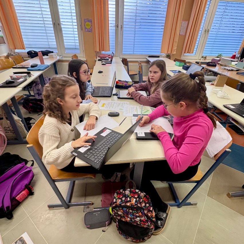 Four young girls in a classroom are working together on laptops and papers. They are surrounded by other students at their desks, with backpacks and books scattered around.