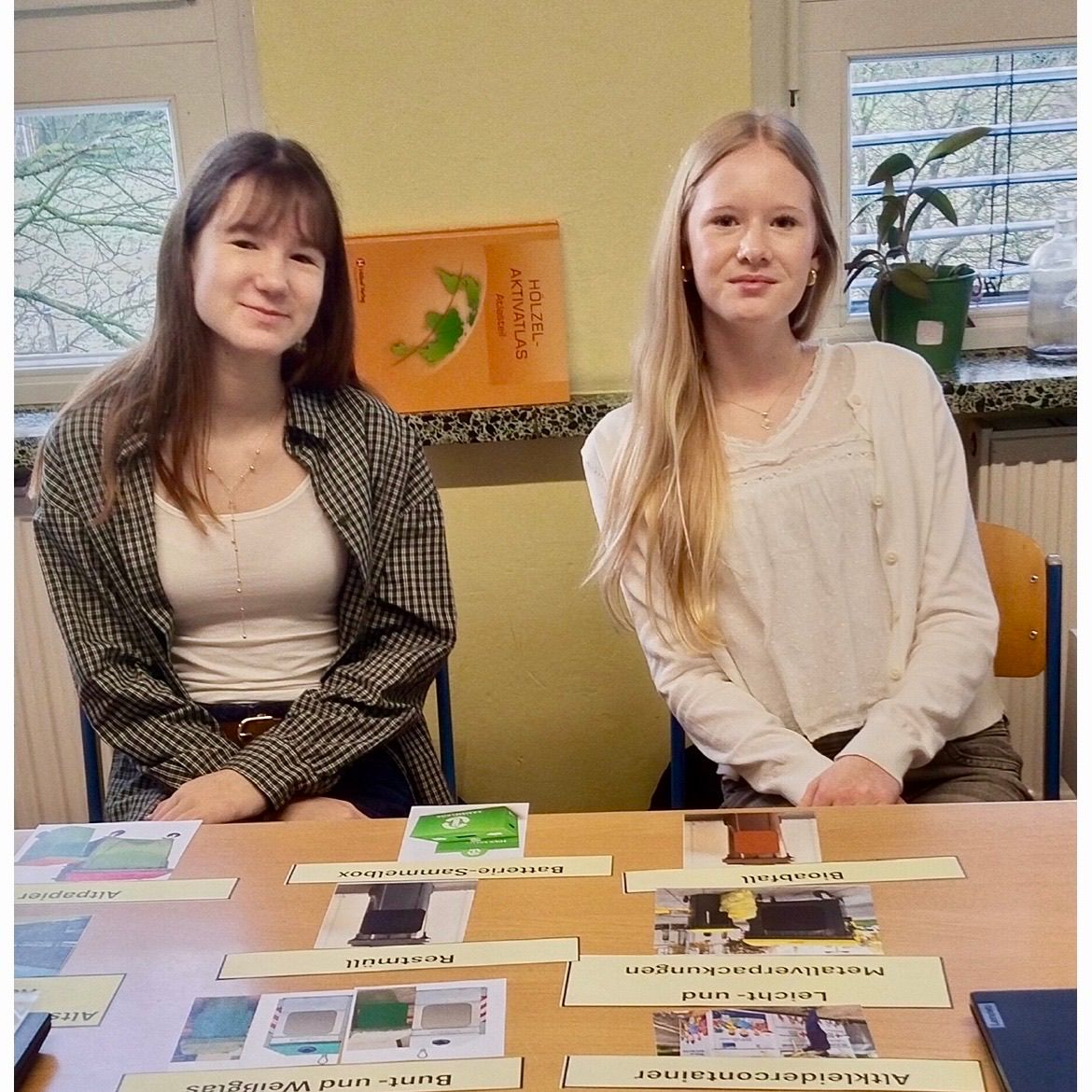 Two women sit at a table with various cards. One wears a checkered jacket, the other a white blouse. Behind them is a yellow wall with a book titled 'Aktivitas' and a potted plant. Windows show trees outside.