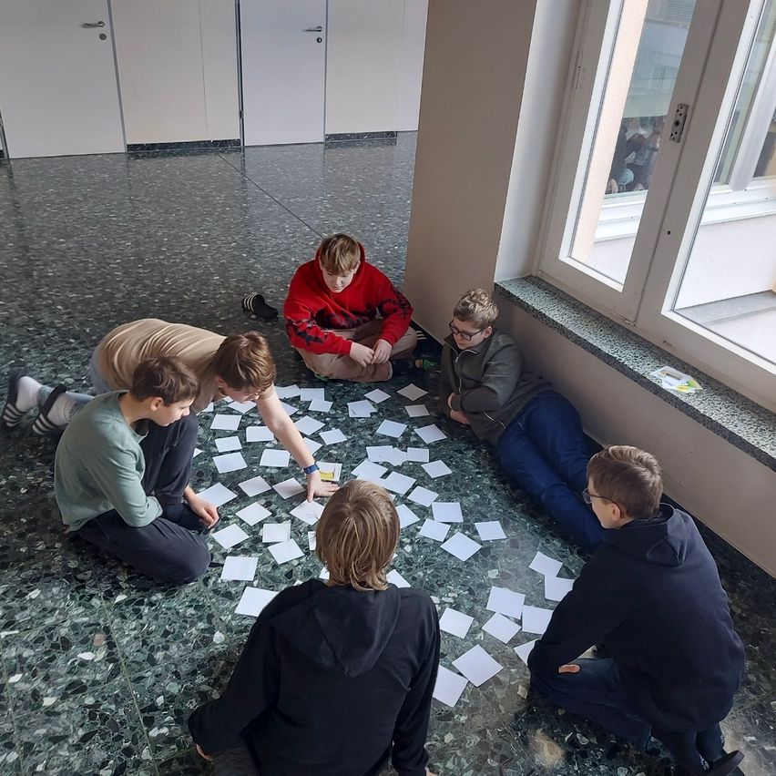 A group of young boys are sitting in a circle on the floor with papers around them, possibly engaged in a group activity.