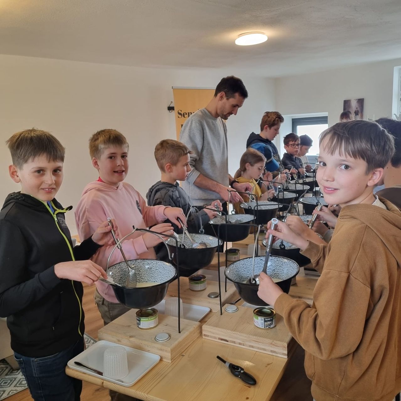 Kinder nehmen in geschlossenen Räumen an einer Kochaktivität teil, jeder mit einem Topf auf einem Holztisch, unter Anleitung eines Erwachsenen.