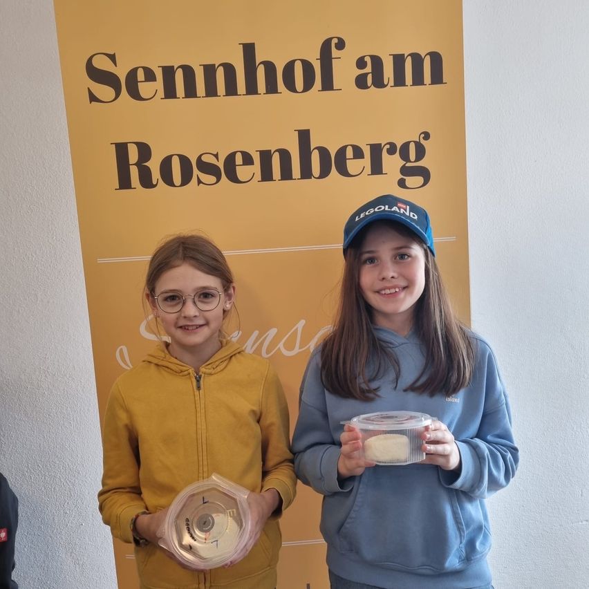 Zwei junge Mädchen stehen vor einem Schild für Sennhof am Rosenberg. Eine hält eine Trophäe und die andere einen Behälter.