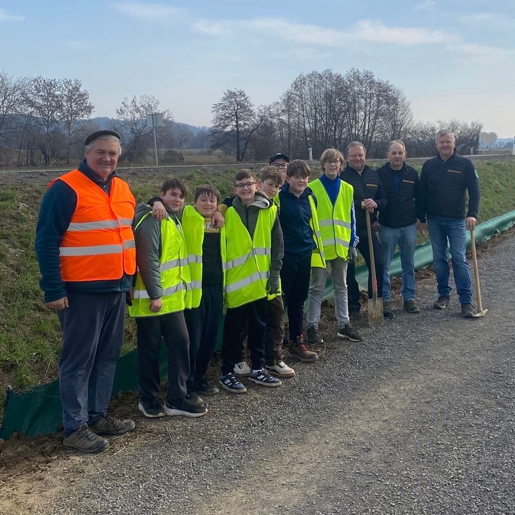 Eine Gruppe von Menschen, einschließlich Kindern, steht mit Warnwesten auf einem Kiesweg, lächelt und posiert für ein Foto. Sie halten Schaufeln und stehen in der Nähe eines Grasfeldes.