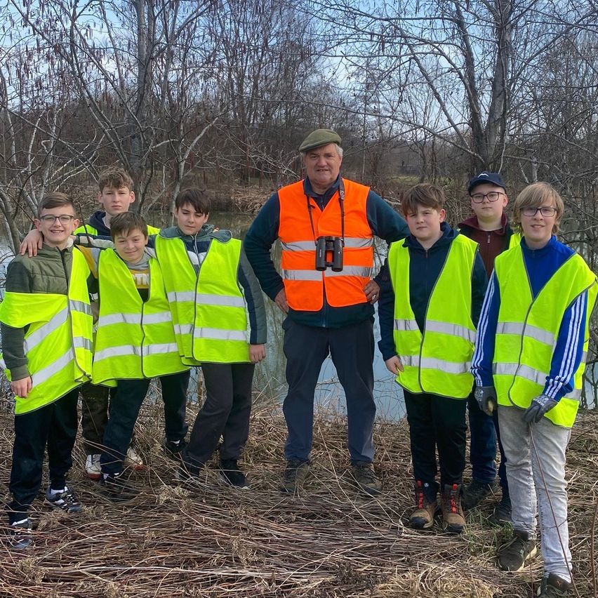 Eine Gruppe von Jungen in Warnwesten und einem Mann mit Fernglas steht am Ufer eines Flusses. Der Mann hat seinen Arm um die Schulter eines Jungen gelegt.